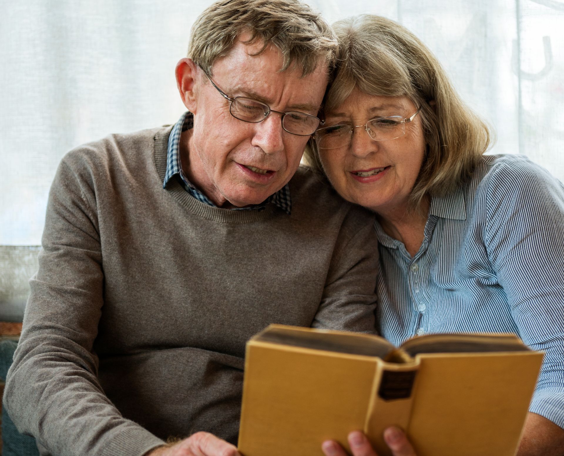 Two people sit close together reading a book, smiling in a cozy indoor setting.