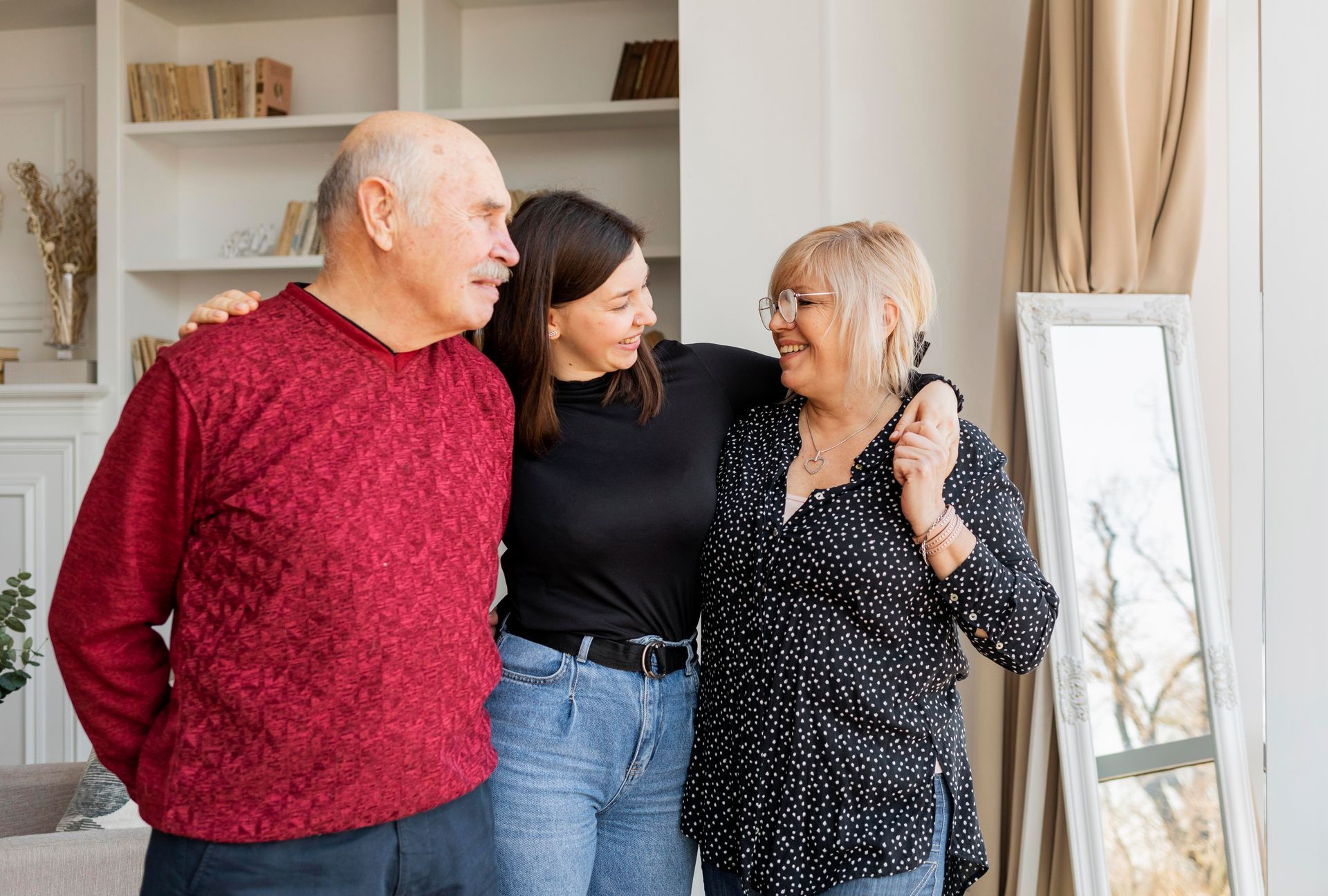 Three people smiling together in a bright living room near a mirror and shelving unit