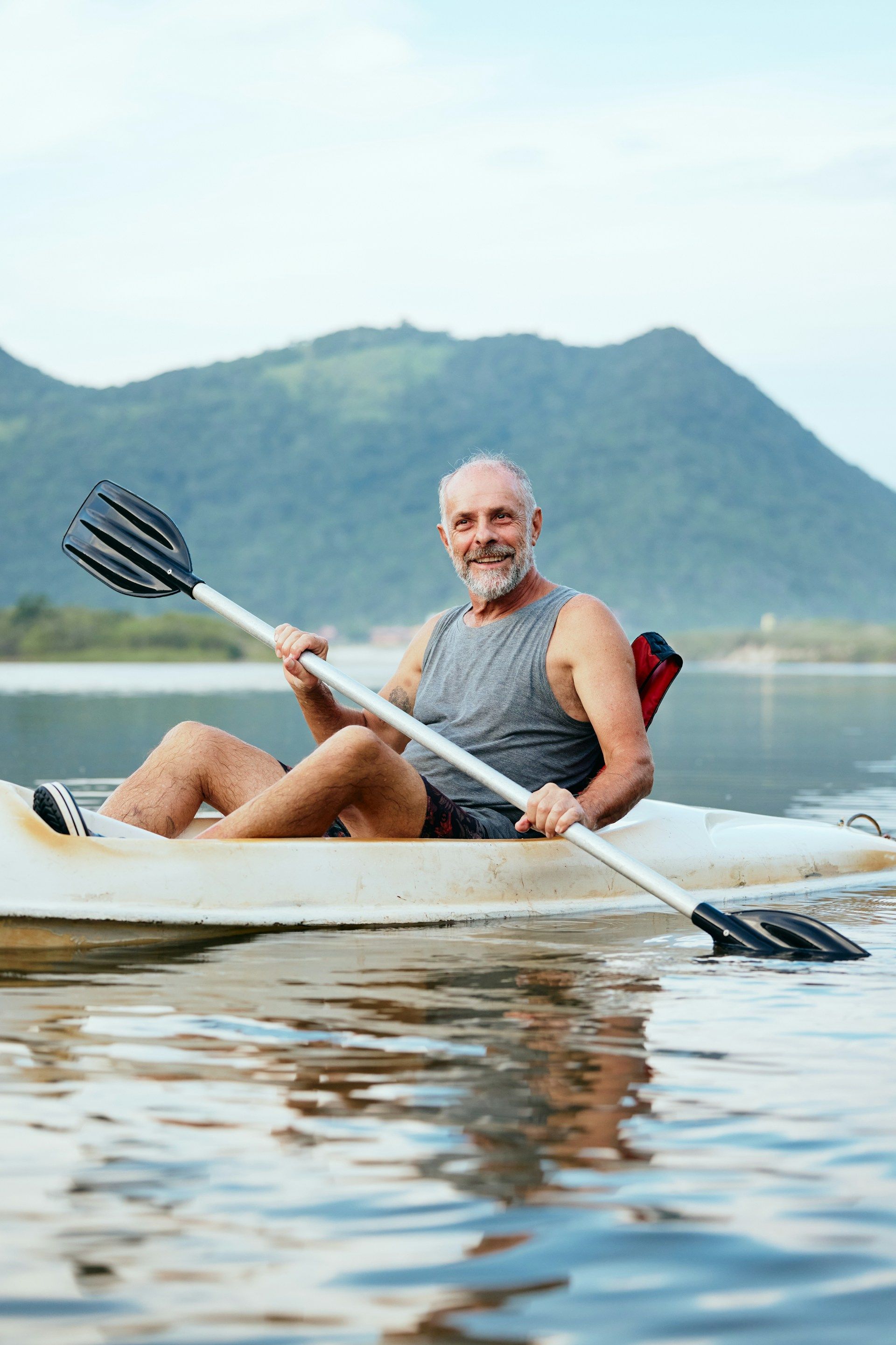 A person kayaking on calm water with a mountain backdrop, smiling and holding a paddle