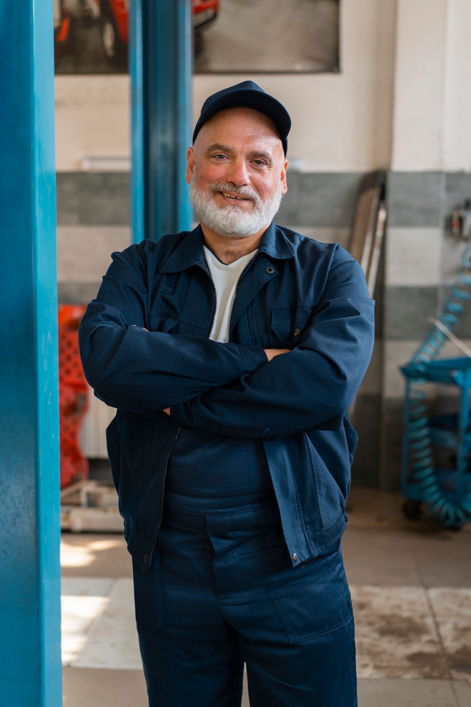 Man standing with arms crossed, wearing a cap and navy jacket in an indoor workshop setting