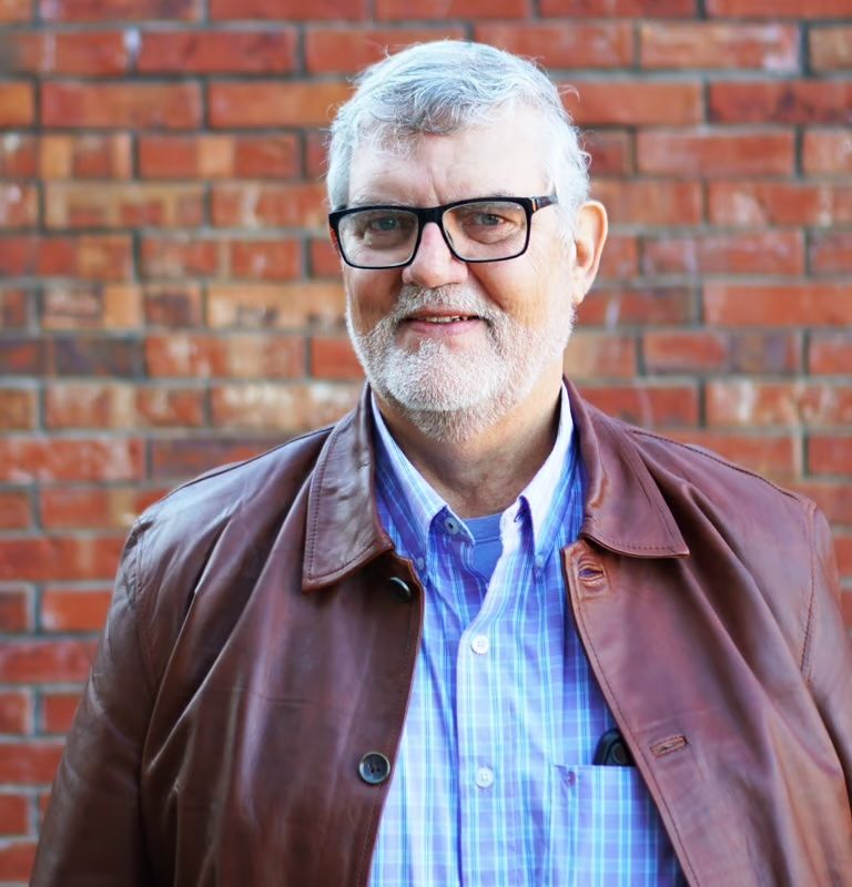 Smiling man with white hair and glasses in a brown jacket, standing against a brick wall.