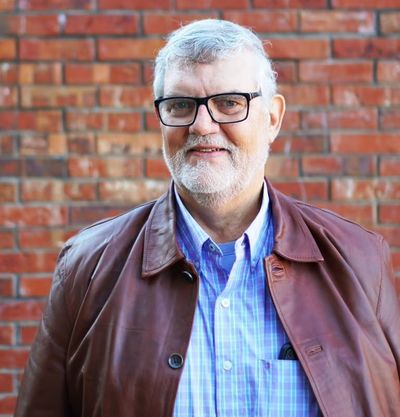 Smiling man with glasses and white hair in a brown jacket, standing against a brick wall.