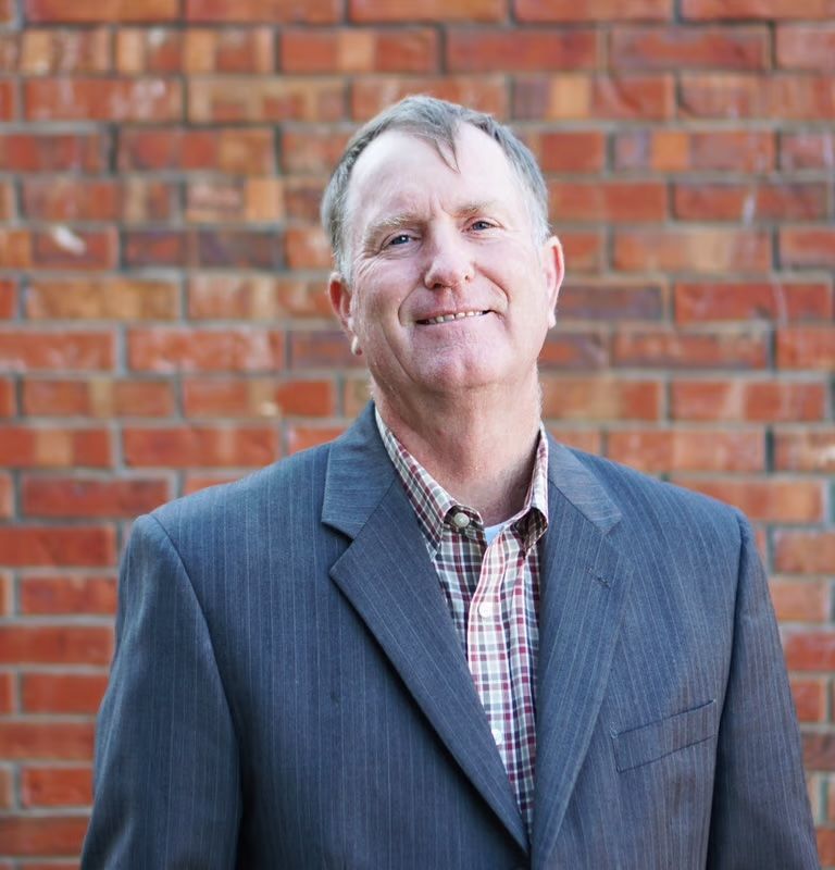Man in gray blazer and plaid shirt smiling in front of a brick wall