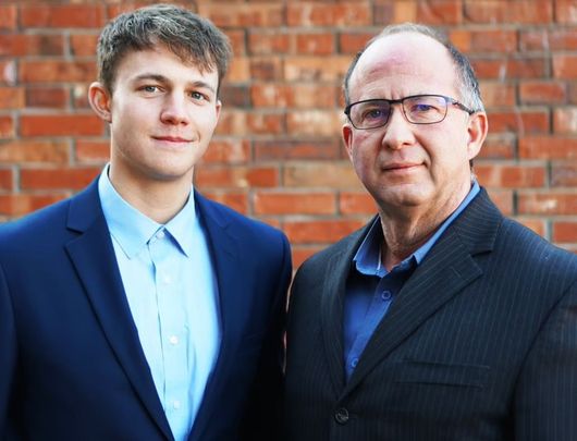 Two men in suits standing in front of a brick wall, one smiling slightly.