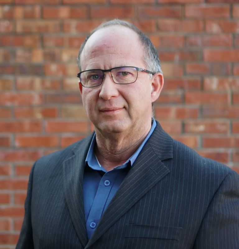 Man in glasses and dark blazer standing before a brick wall