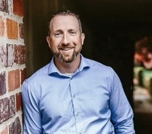 Smiling man in a blue shirt leaning against a brick wall in a doorway