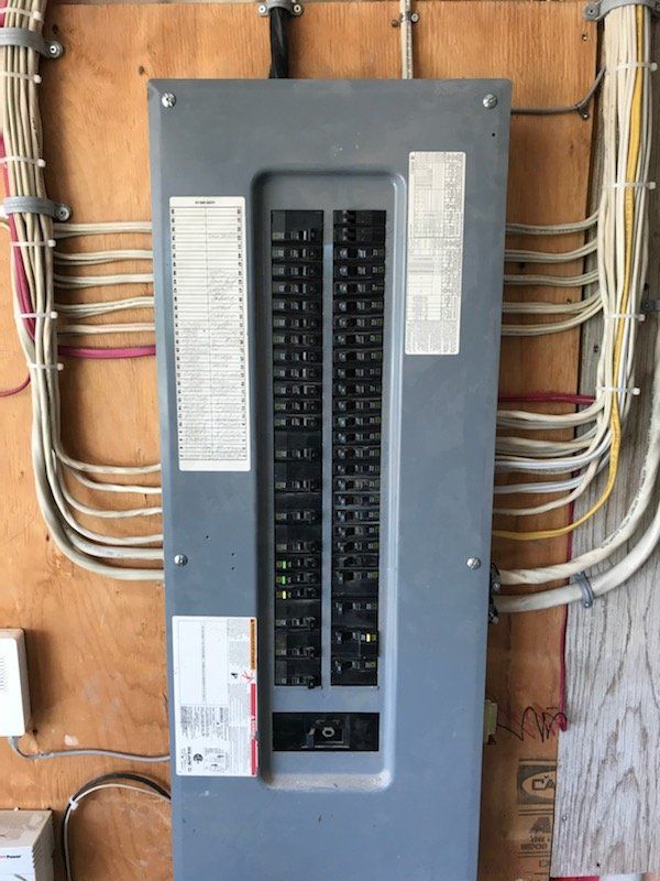 Panneau électrique avec disjoncteurs multiples, couvercle gris et circuits étiquetés sur panneaux de bois.