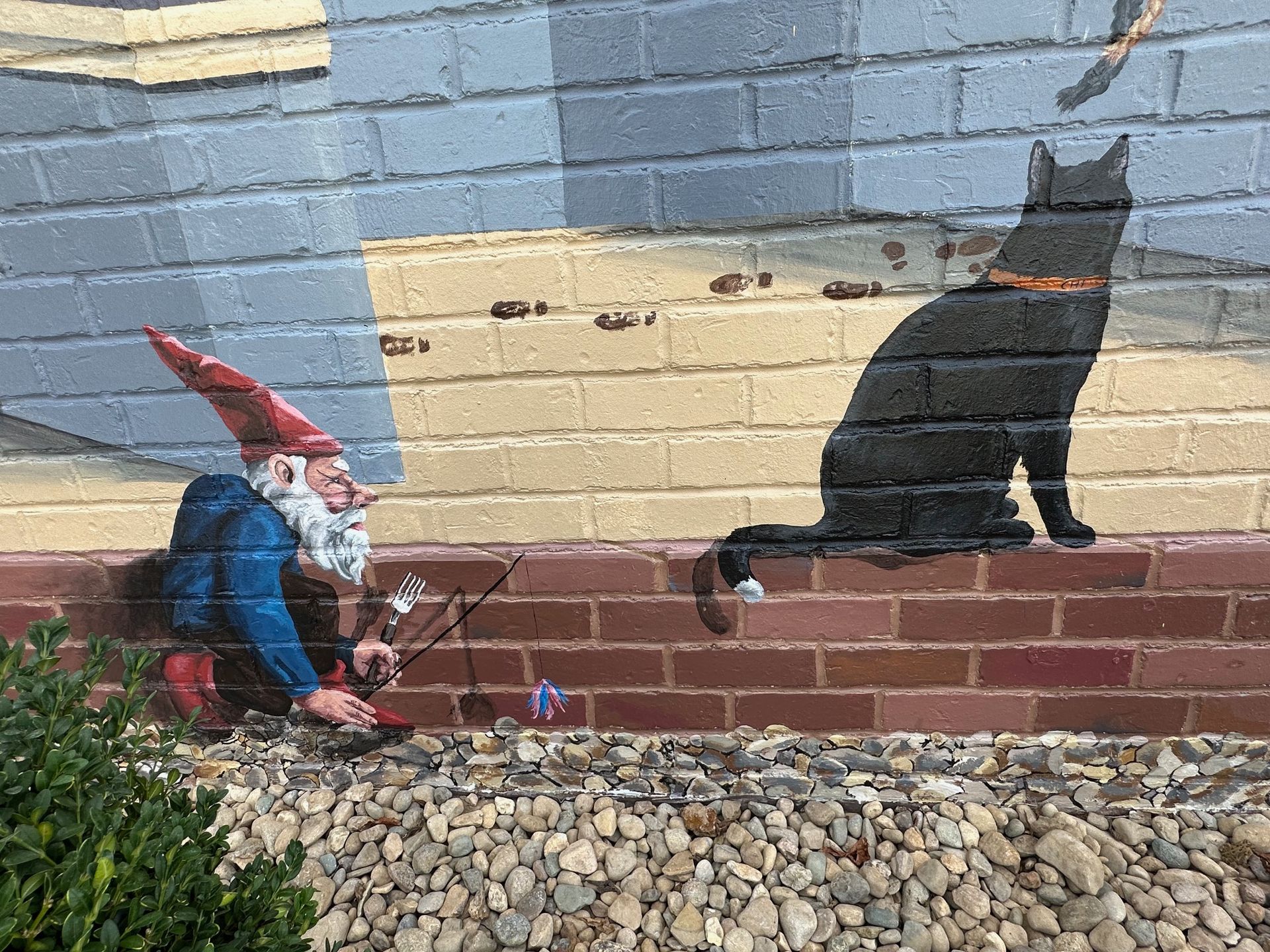 A gnome is playing a violin next to a black cat on a brick wall.