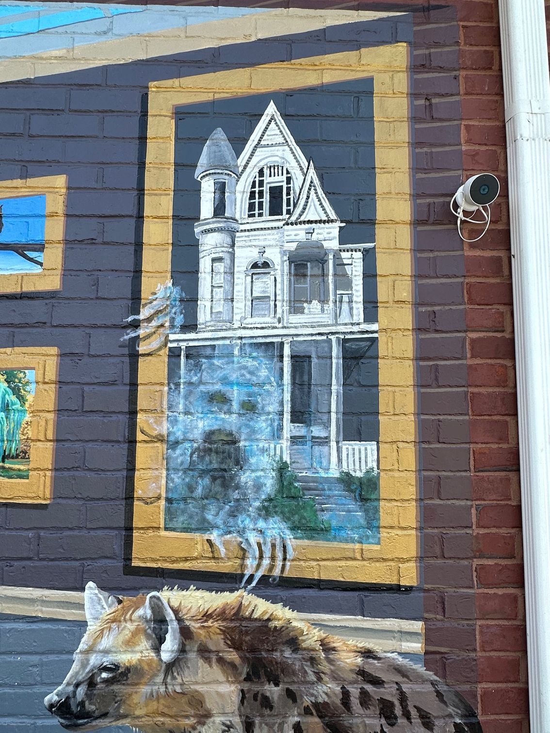 A hyena is standing in front of a painting of a house on a brick wall.