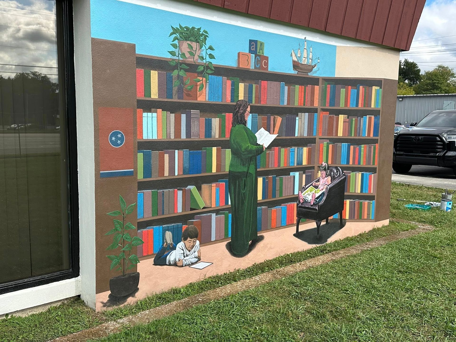 Mural of a library scene: person reading in green robe, child reading, bookshelves, and plants.
