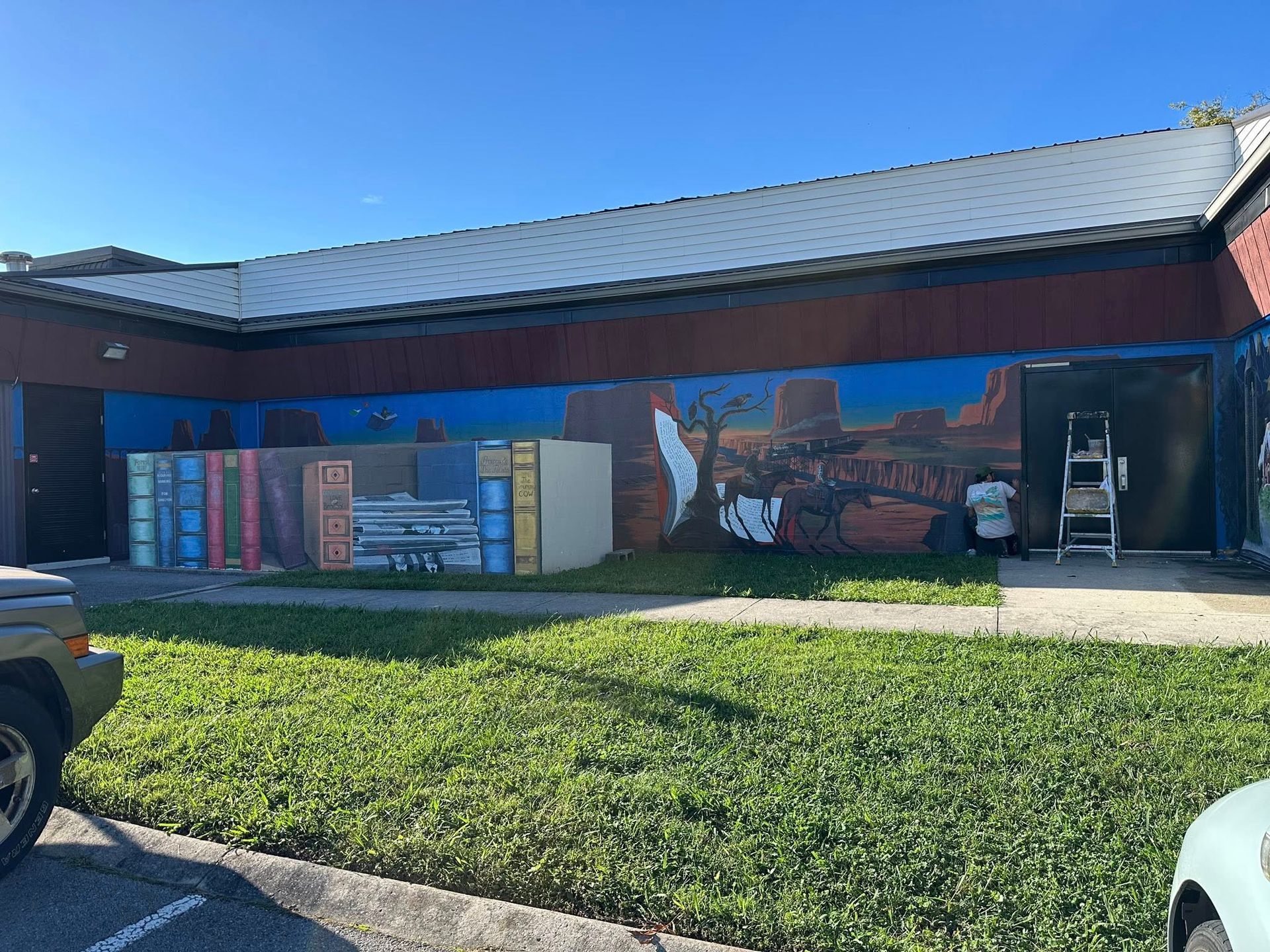 Mural on building exterior: Books, landscape scene, and a ladder against the wall in daylight.