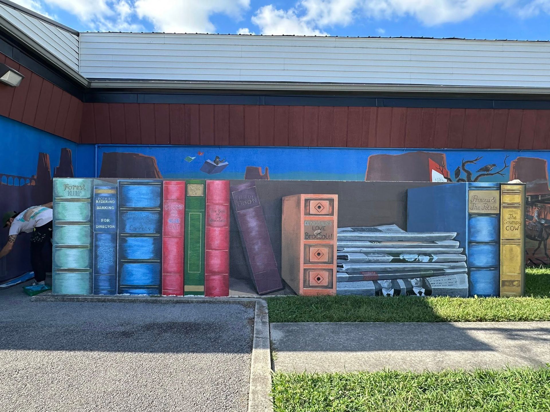 Mural of large books on a building's wall with a person tending to the painting.