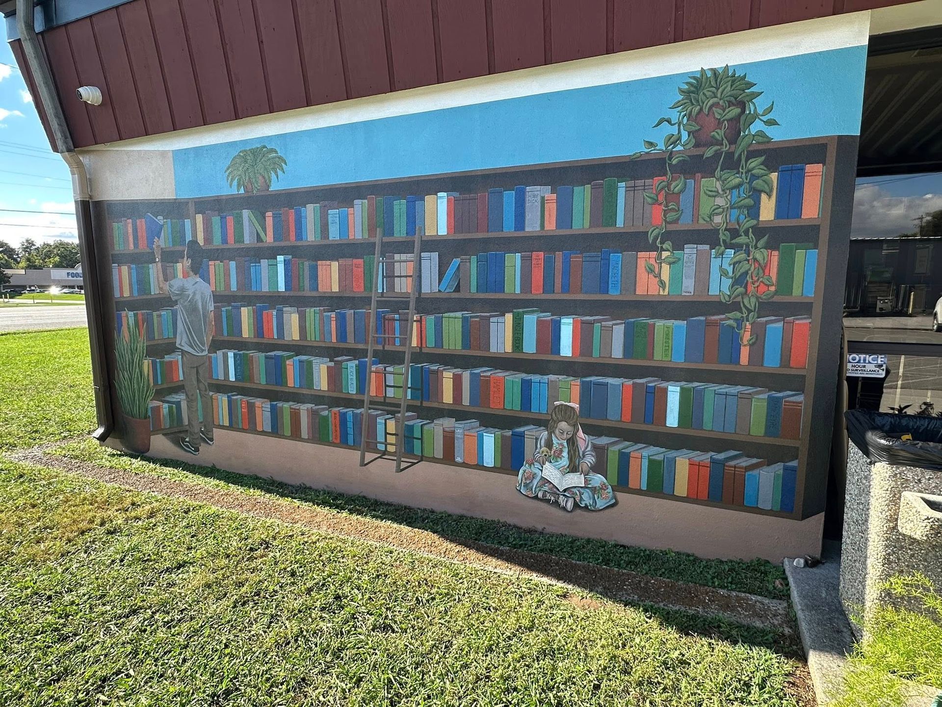 Mural on a building depicts a large bookshelf with a person on a ladder, reaching for books.