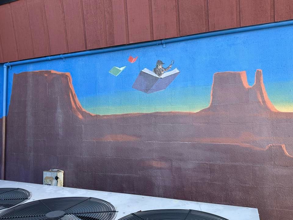 Mural of person flying on a book over a desert landscape with mesas. Blue sky, colorful.