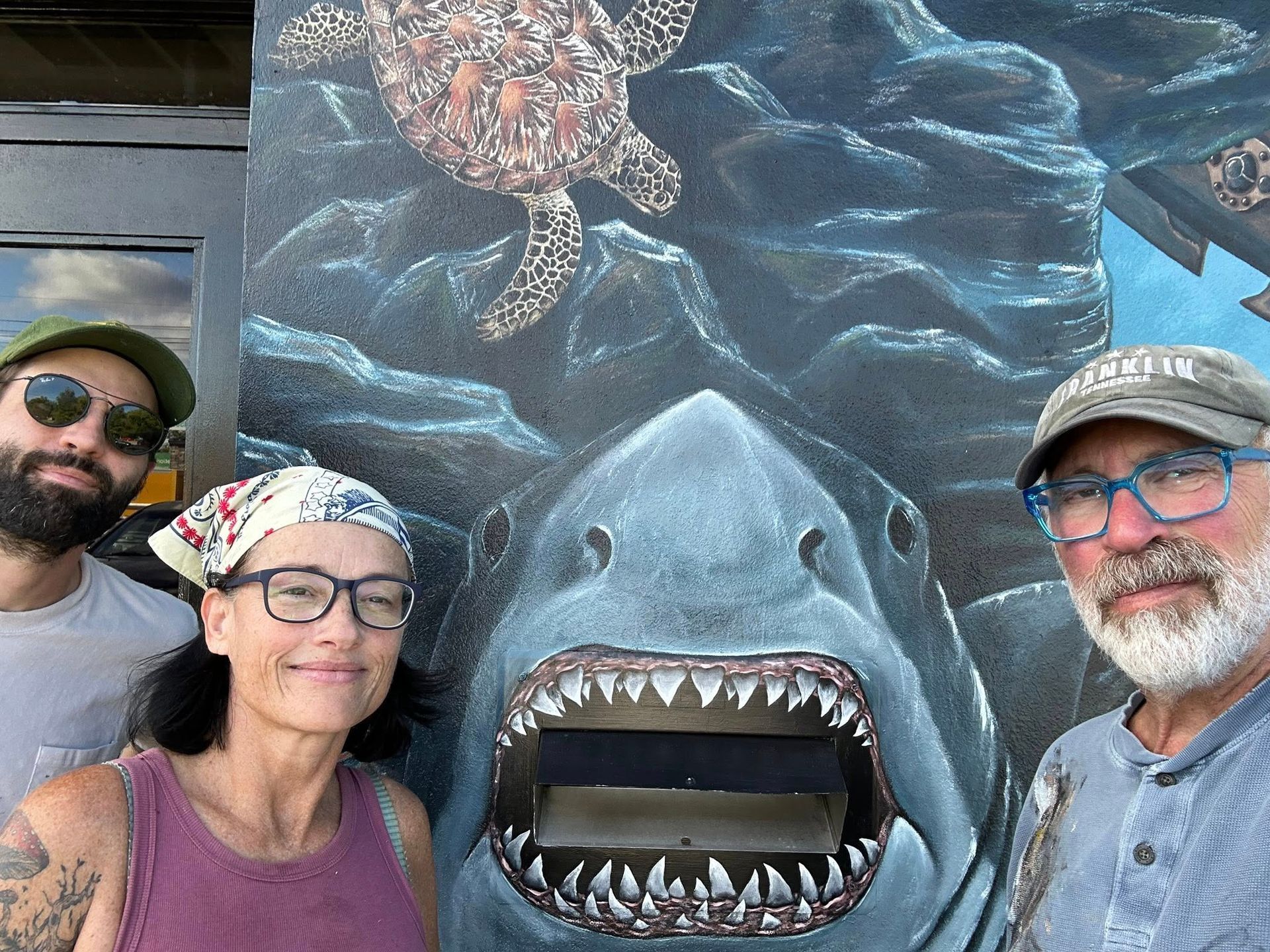 Three people pose with a large mural of a shark and sea turtles. The shark has an open mouth, surrounded by the ocean and rocks.