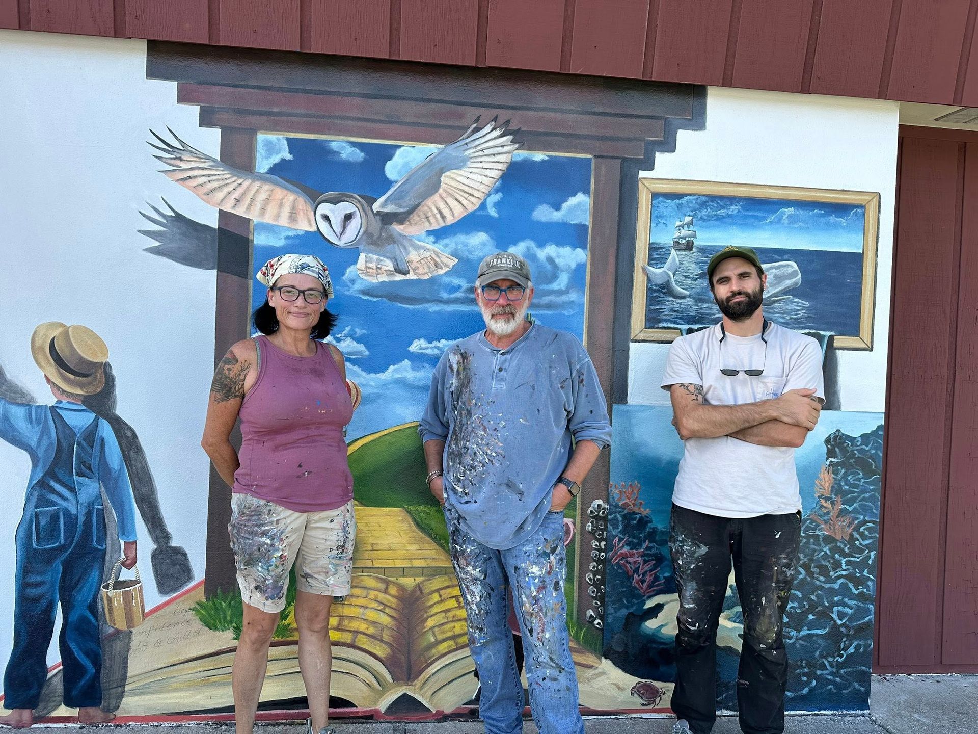 Three people stand in front of a colorful mural of an owl, landscape, and figures. All are covered in paint.