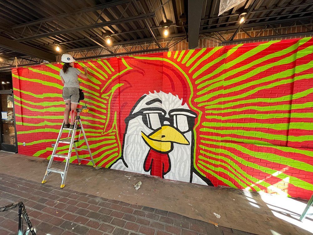 A woman is standing on a ladder painting a chicken mural on a wall.