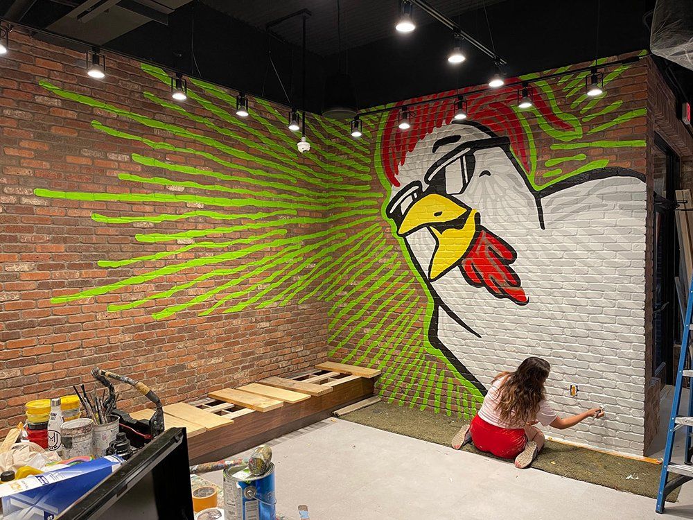 A woman is painting a mural of a chicken on a brick wall.