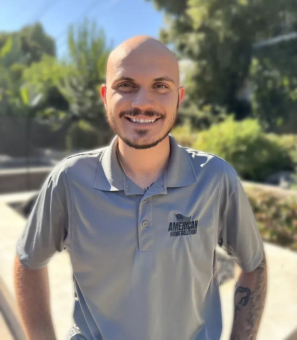 Smiling bald man in gray polo shirt standing outdoors in bright sunlight.