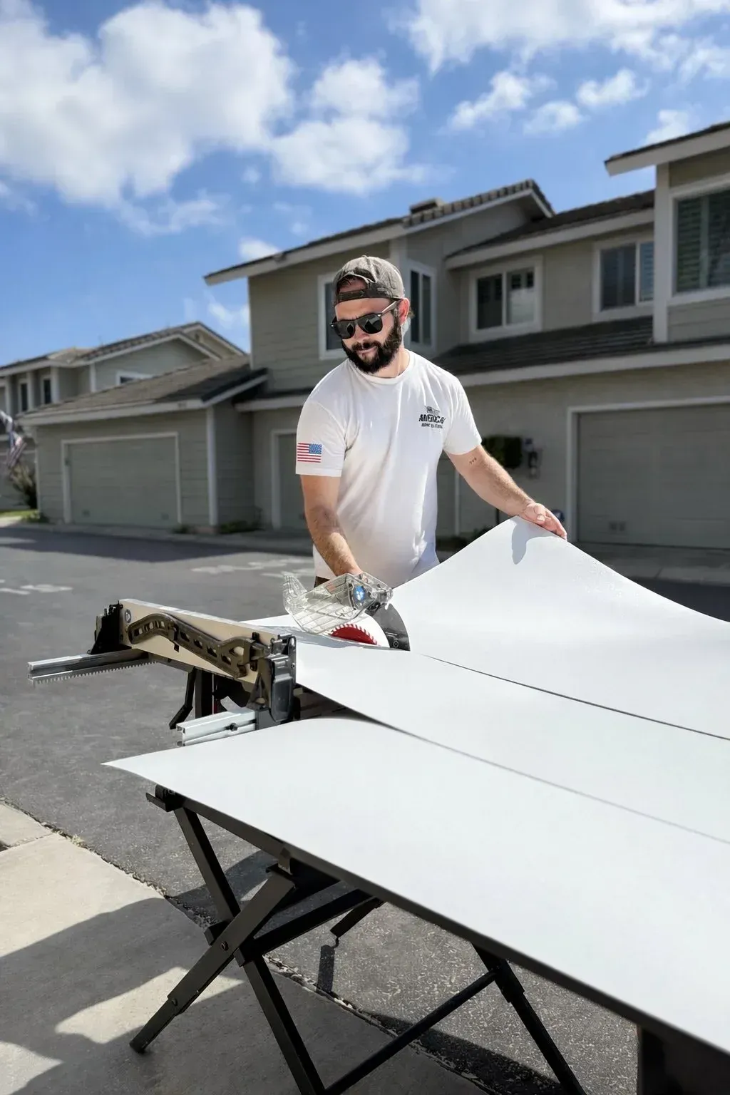 Man cutting a white board outdoors beside suburban garages under a blue sky