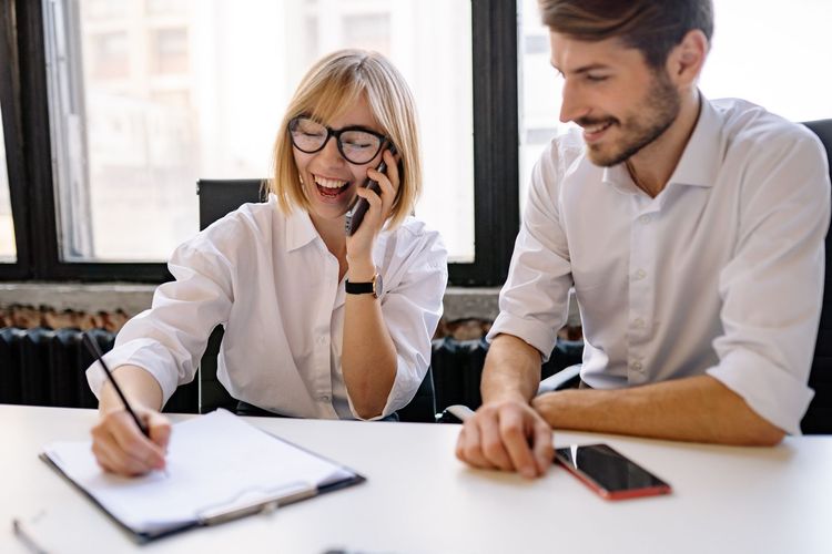 Two coworkers smiling at a desk, one writing in a notebook while the other points beside a smartphone.