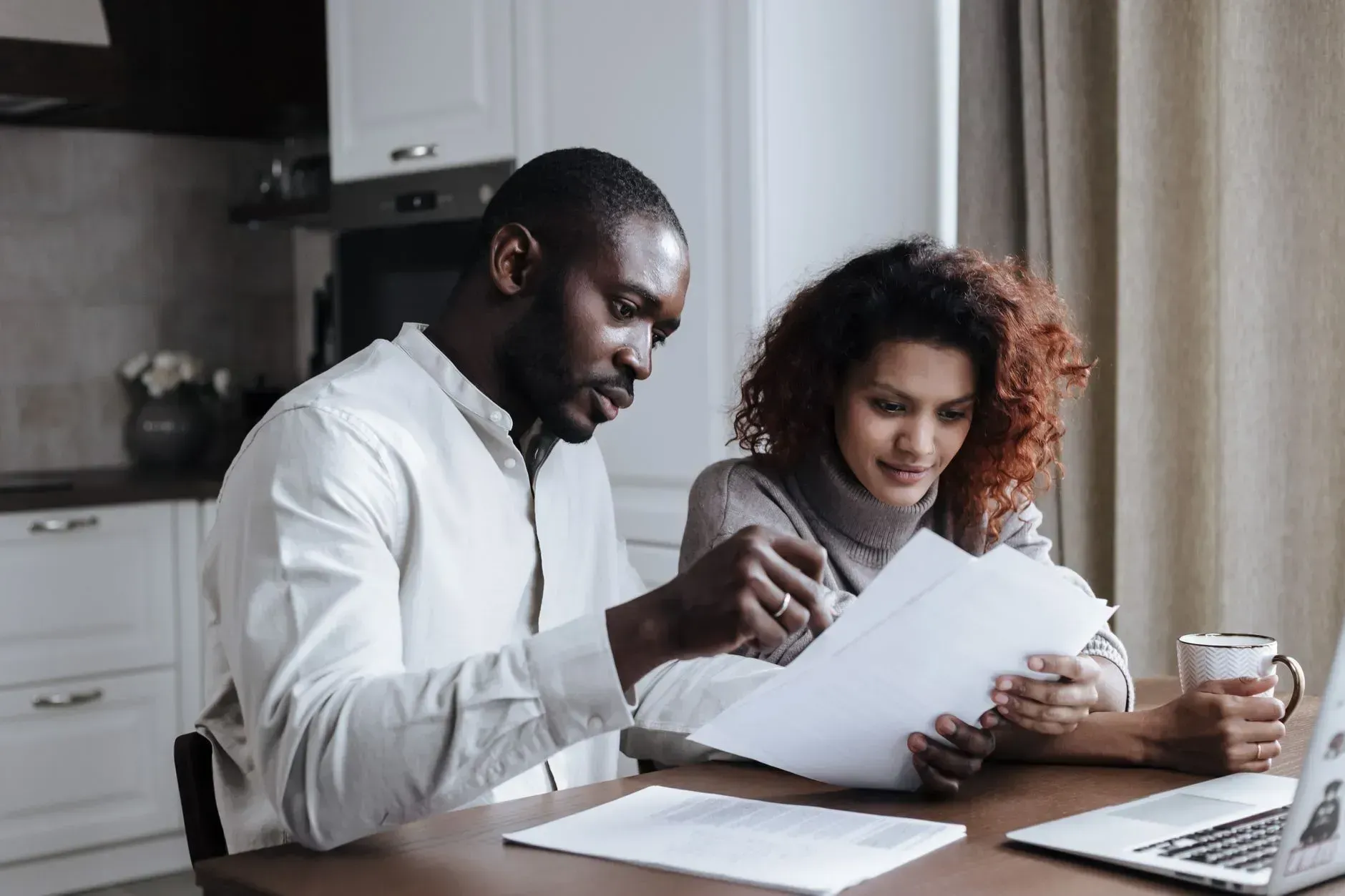 Two people reviewing paperwork together at a kitchen table with a laptop and coffee mugs.