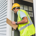 Delivery worker in a yellow safety vest and hard hat checking a clipboard at a front door