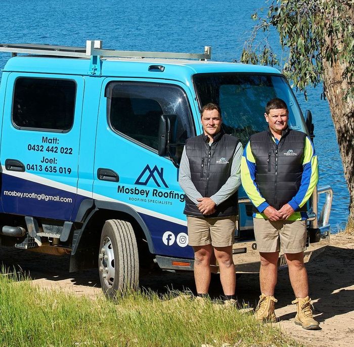 Two men standing in front of a blue Mosbey Roofing work truck by a body of water.