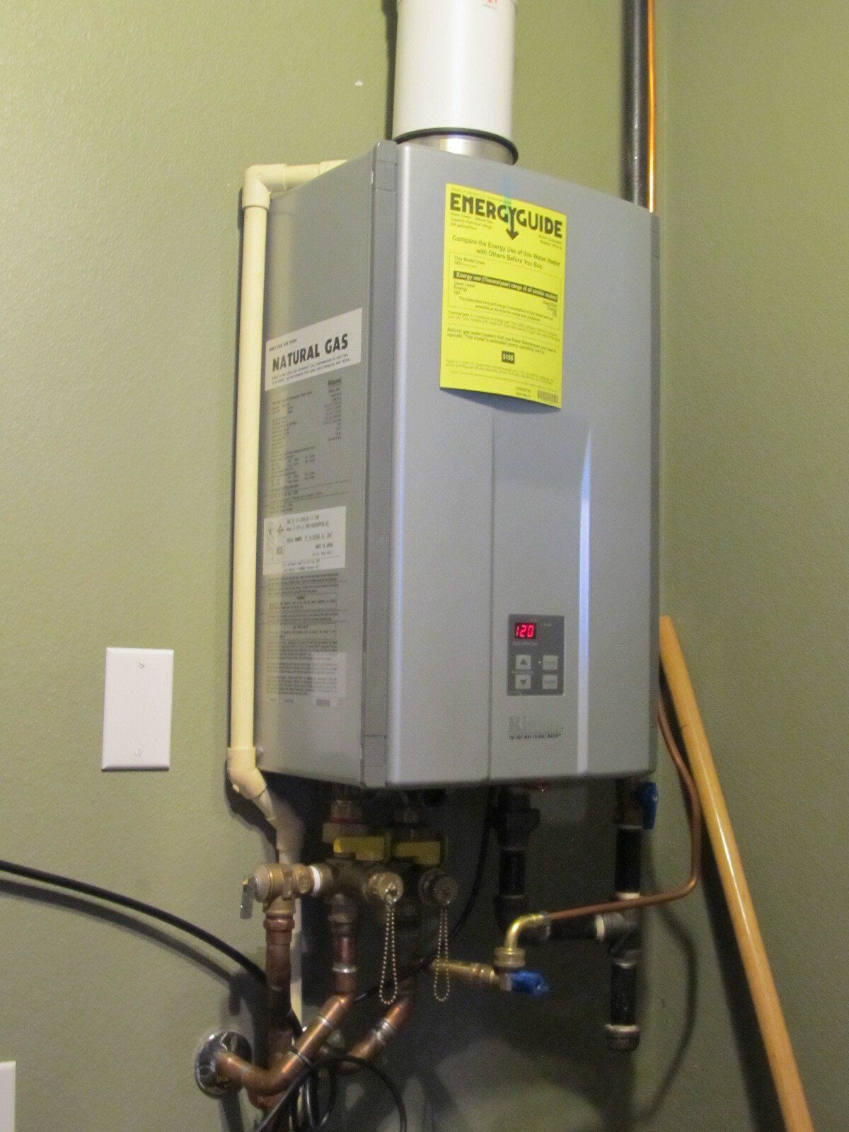 Wall-mounted tankless water heater with pipes and a yellow service label in a utility room