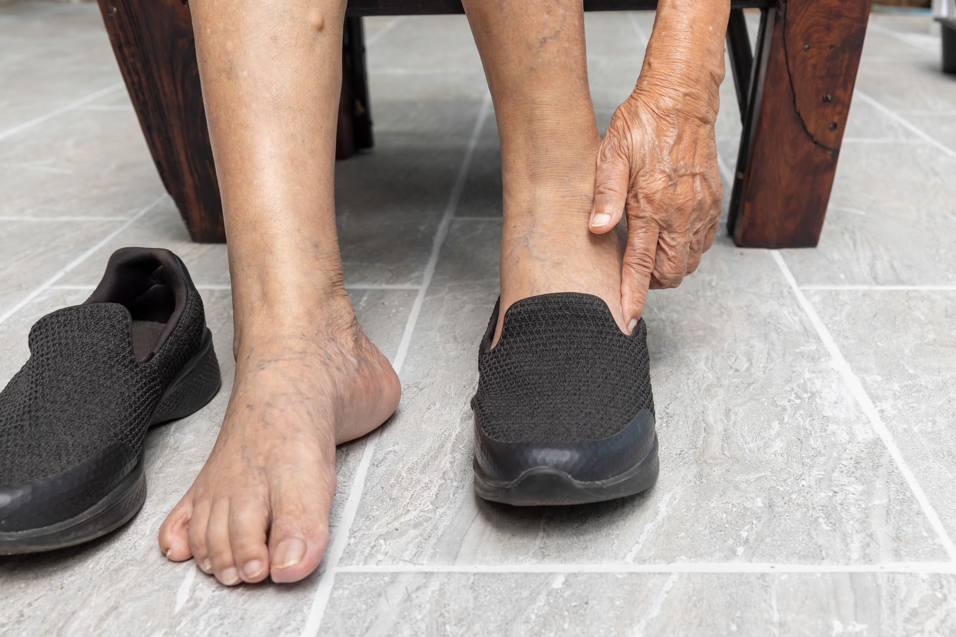Person seated, putting on a black shoe. One bare foot is visible. Gray tiled floor.