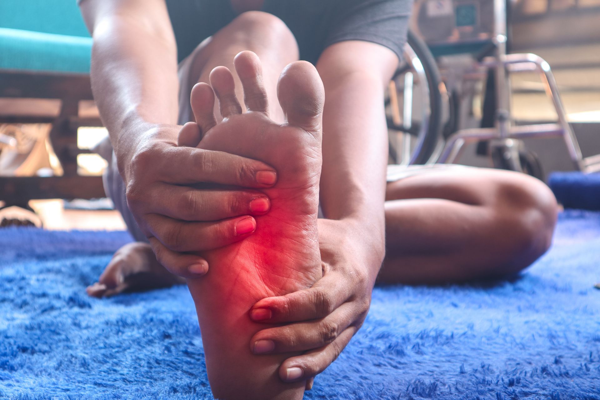 Person seated, holding foot, showing pain area highlighted in red, on blue rug.