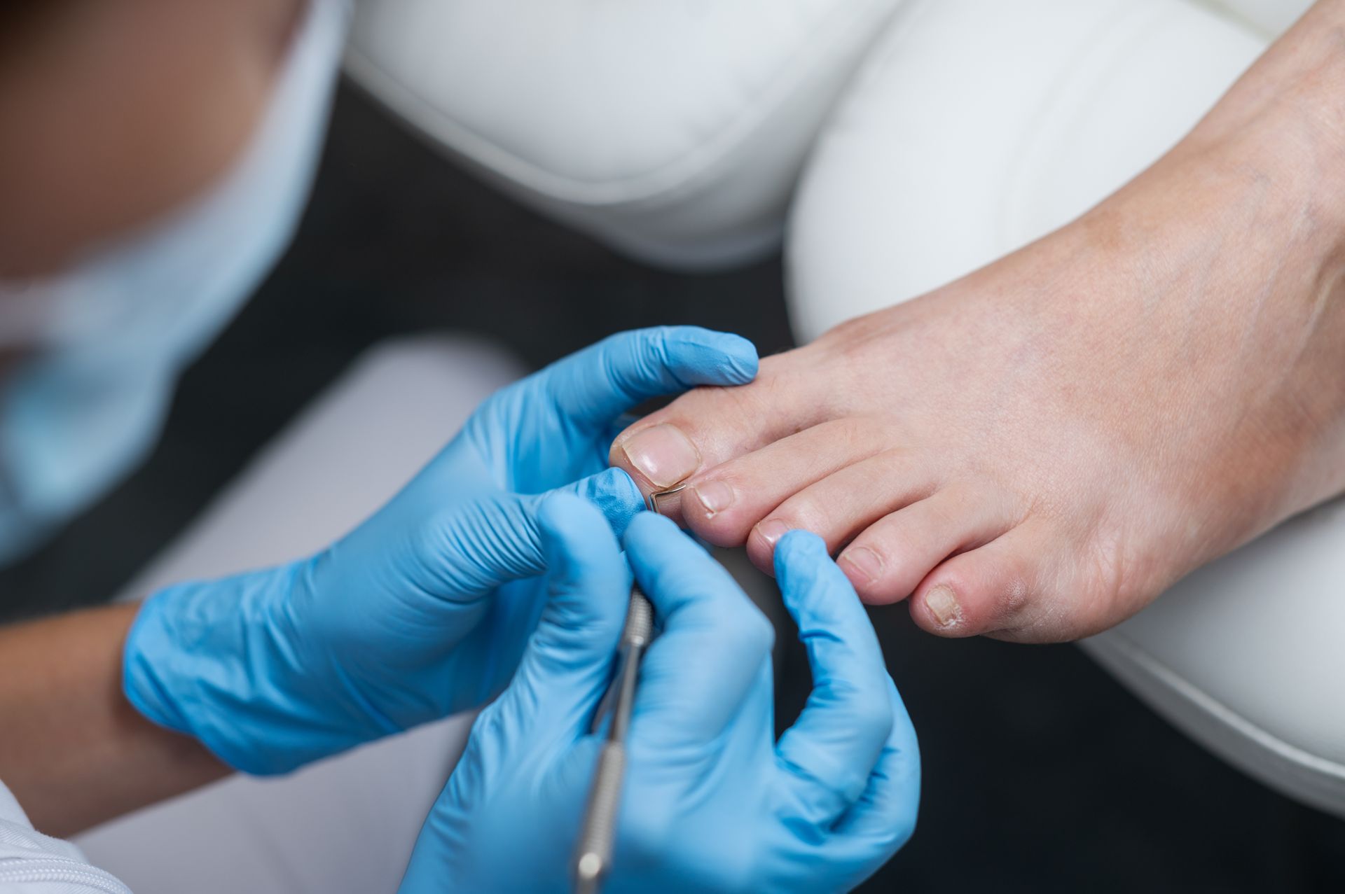 Person's foot receiving a pedicure; gloved hands use a tool to trim toenails. White chair in the background.