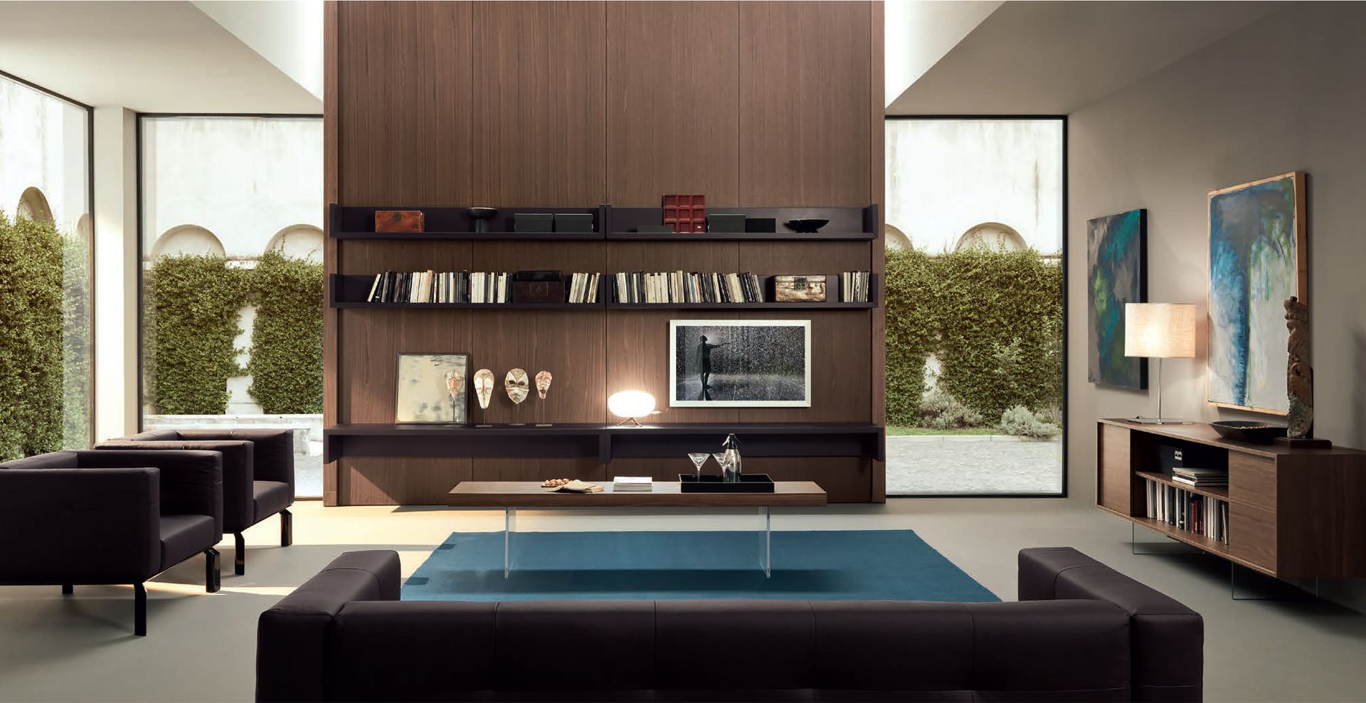Soggiorno moderno con parete attrezzata in legno marrone scuro, tavolino quadrato blu, mobili scuri e ampie finestre con verde.