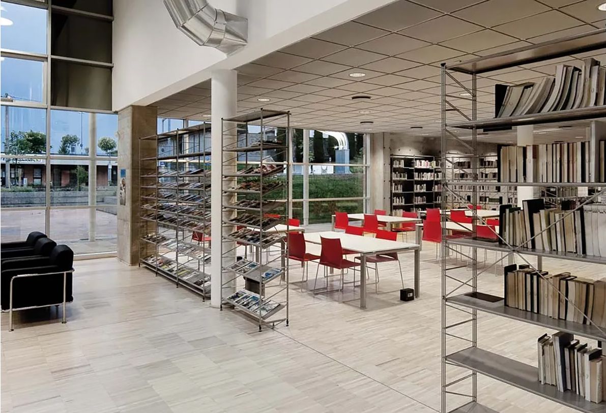 Biblioteca interna con scaffali, tavoli e posti a sedere vicino alle grandi finestre.