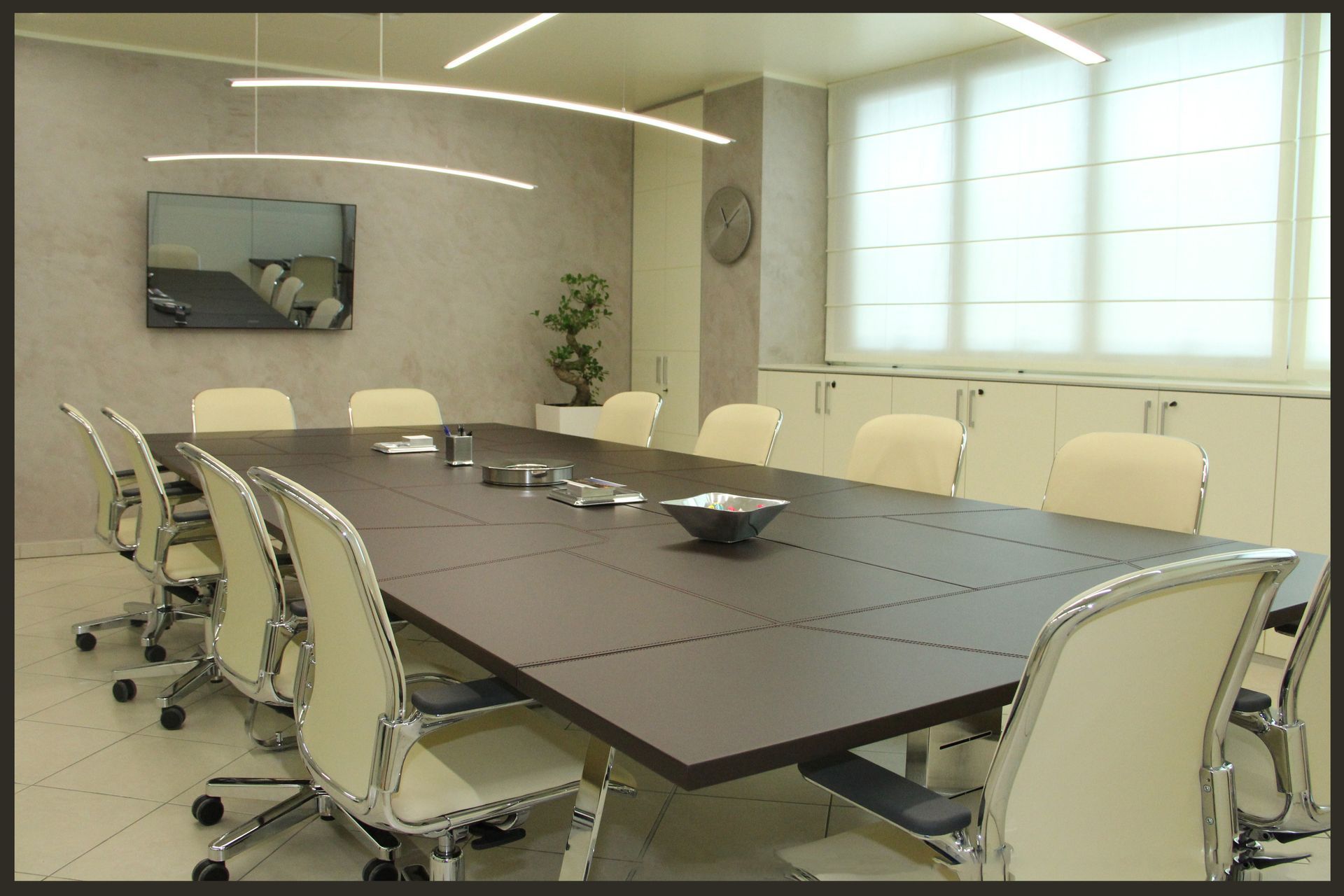 Sala conferenze moderna e vuota con lungo tavolo, sedie color crema, TV a parete e ampie finestre.