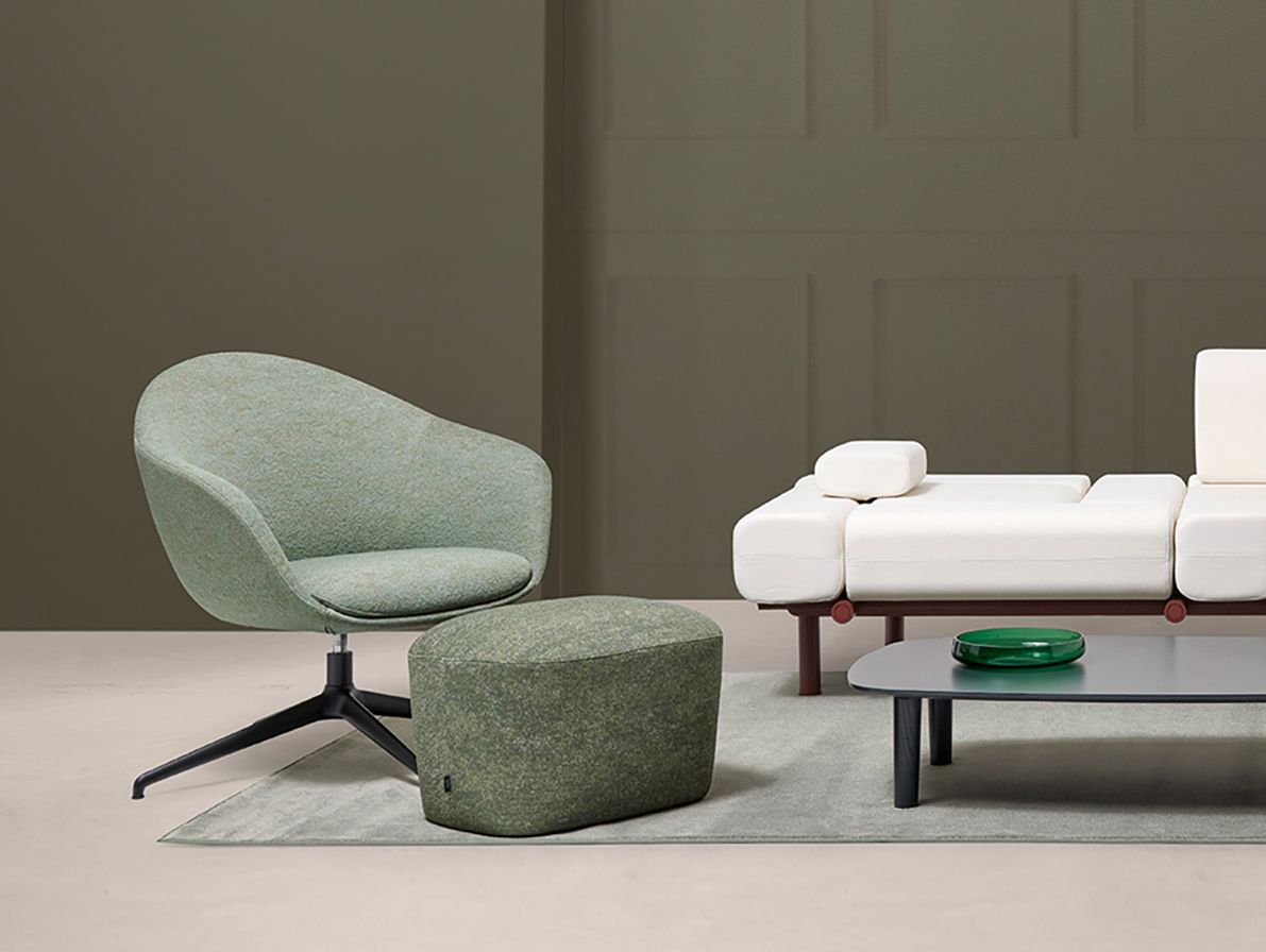 Una moderna sedia verde e un pouf con base nera, accanto a un divano modulare bianco, contro una parete verde tenue.