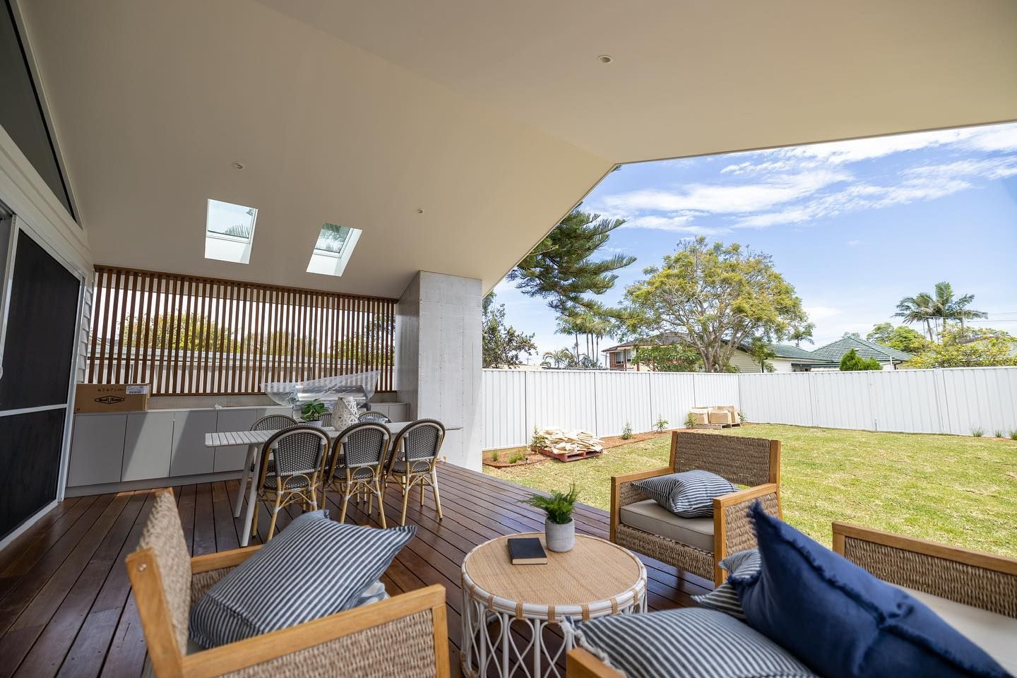 Outdoor deck with wicker furniture, dining table, and a grassy backyard under a bright blue sky. — Blue Horizon Projects in Bateau Bay, NSW