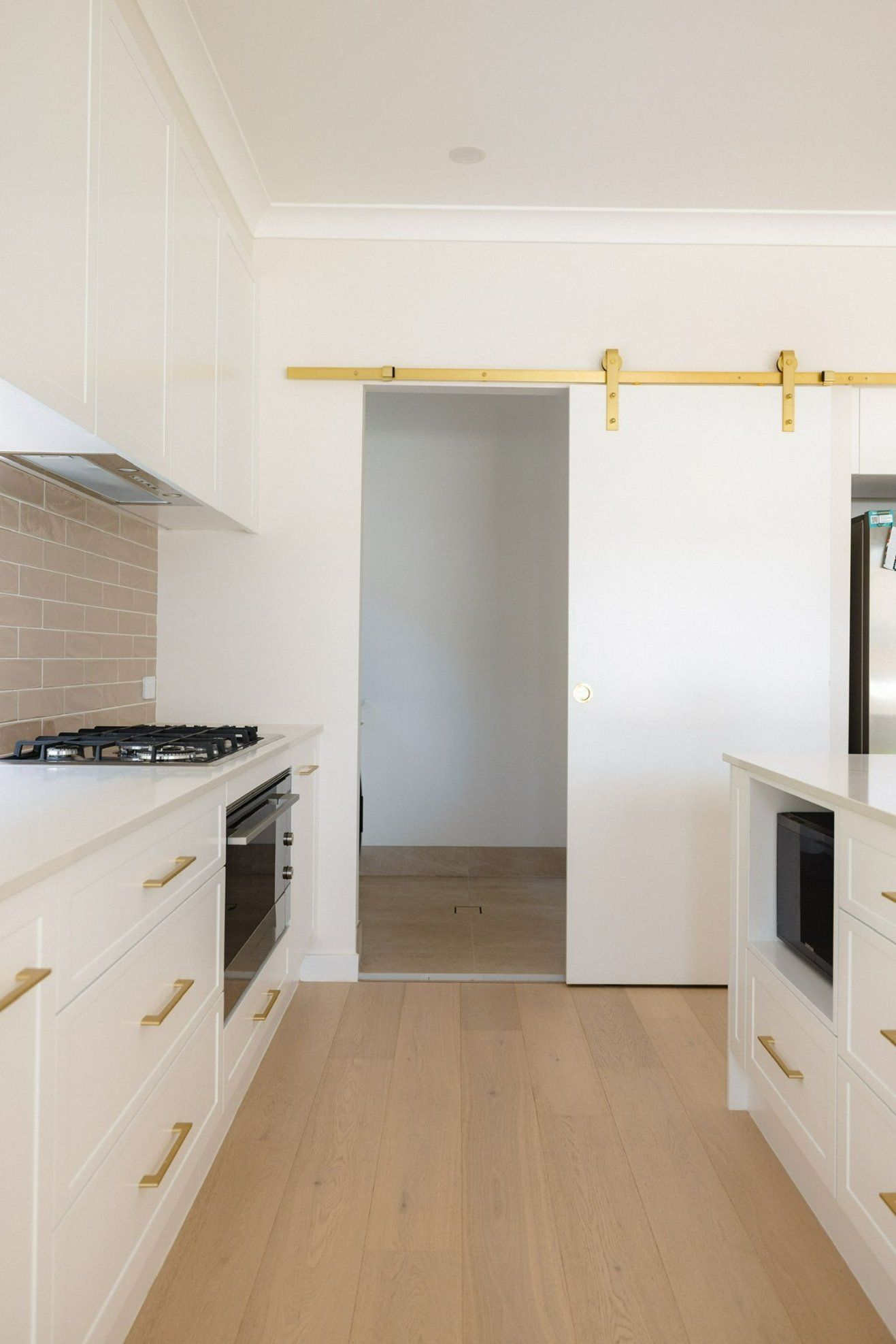 White kitchen with light wood floors and golden hardware; a sliding door leads to another room. — Blue Horizon Projects in Long Jetty, NSW