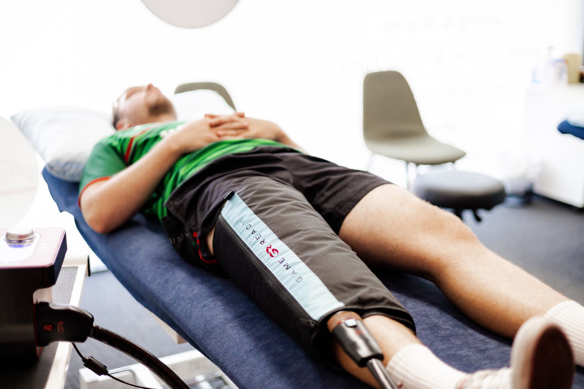 Man lying on a medical table with a leg brace. He's wearing a green shirt and shorts. Light-filled room.