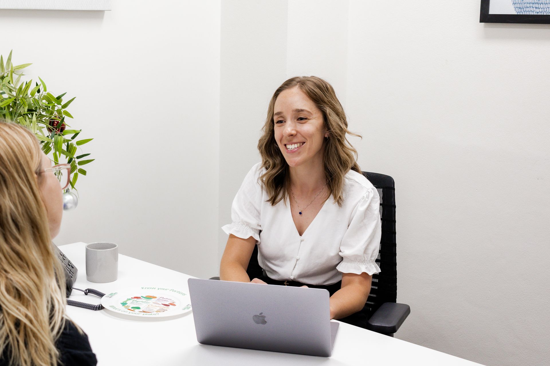 Woman at desk with laptop smiling at another woman; office setting.