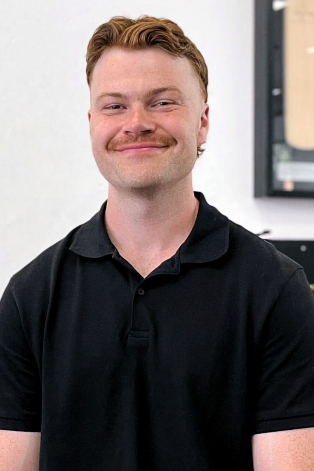 Man with reddish-brown hair and mustache smiles; wearing a black polo shirt. Background is a white wall and a framed picture.