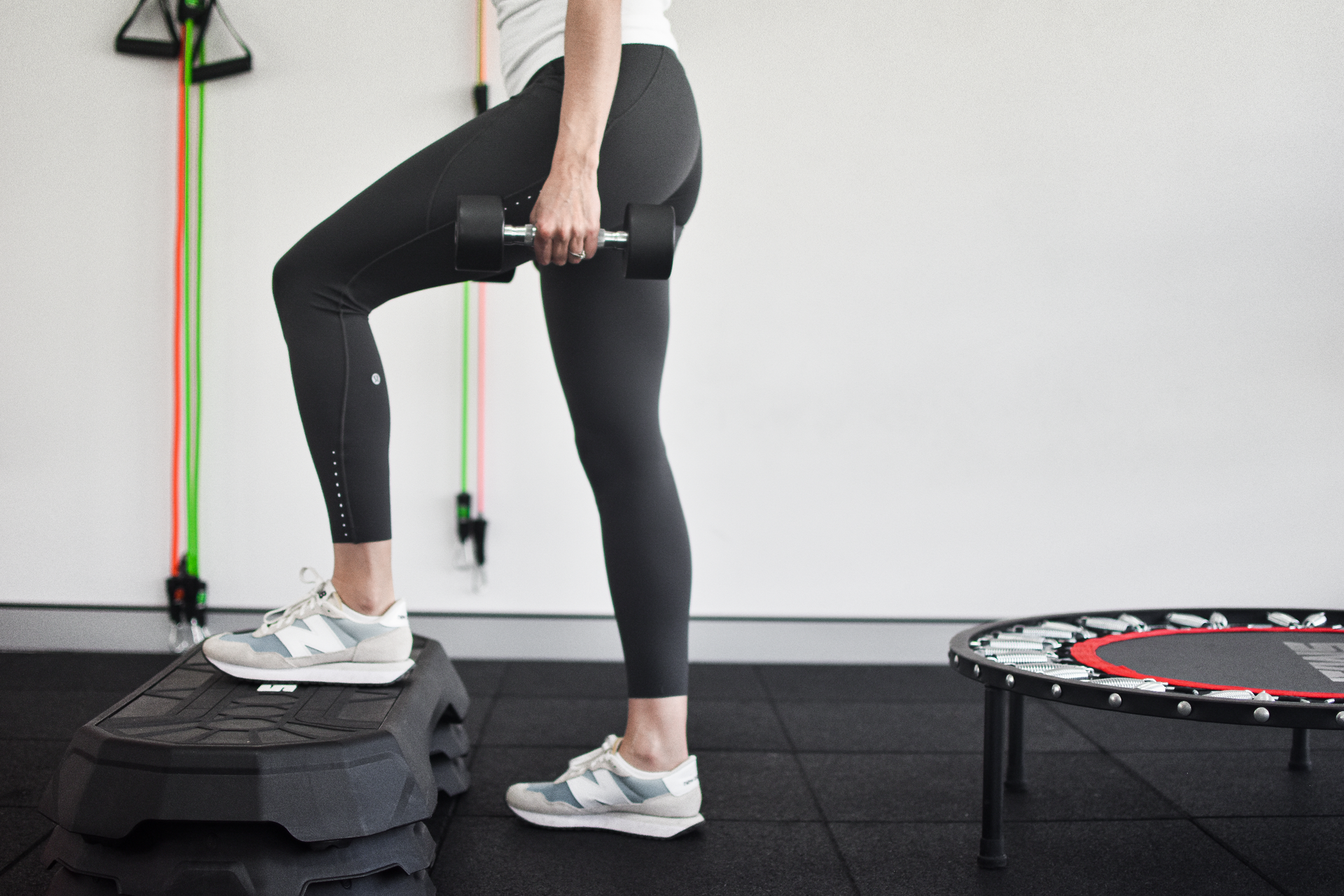 Woman stepping up onto a black step with a dumbbell in hand, near a trampoline.