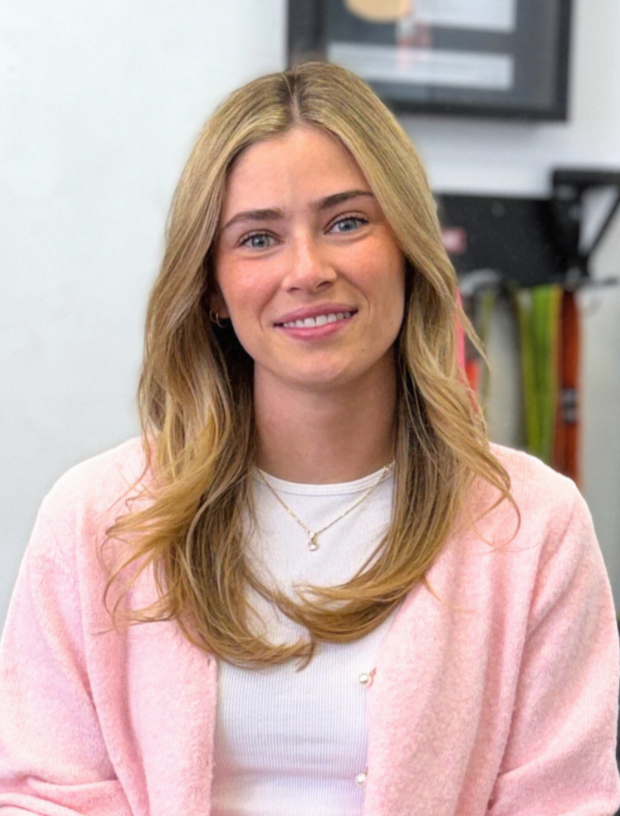Blonde woman in pink cardigan smiles at the camera, necklace visible.