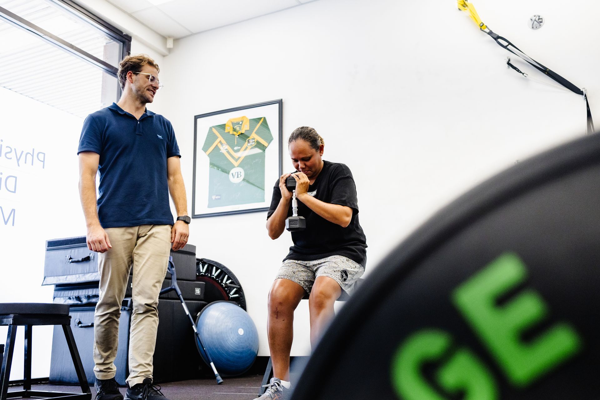 Man watches a person seated, lifting a dumbbell in a gym setting.