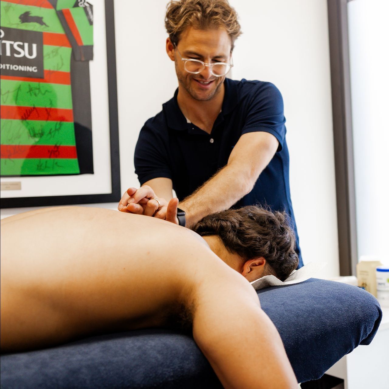 Man giving a back massage to a person lying face down on a massage table; bright office setting.