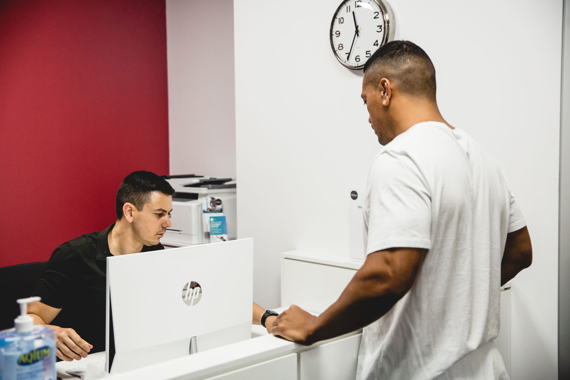 Two men at a reception desk. One man is looking at the computer, while another stands and looks at him.