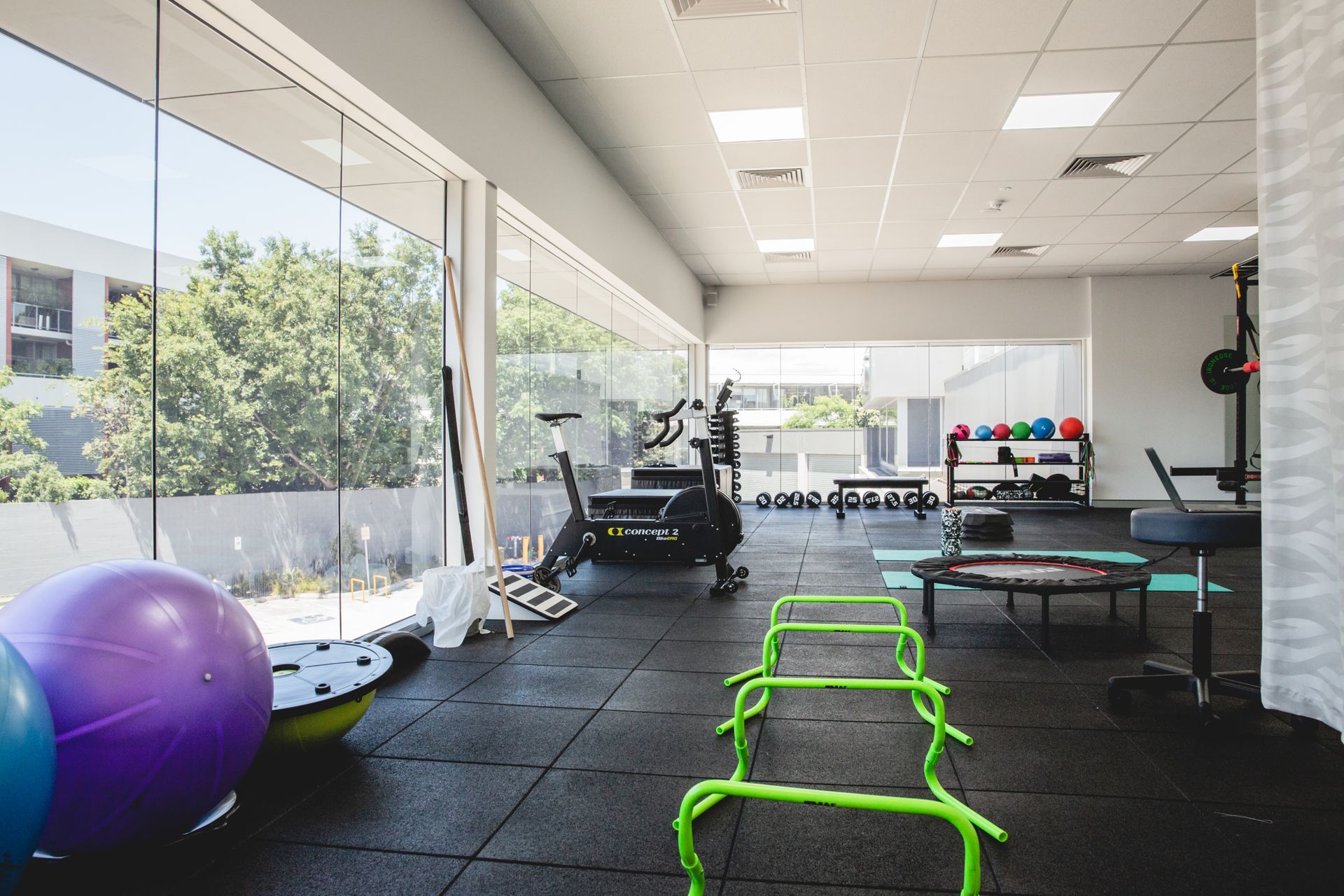 Gym with exercise equipment, including hurdles, stationary bike, and medicine balls, next to large windows.