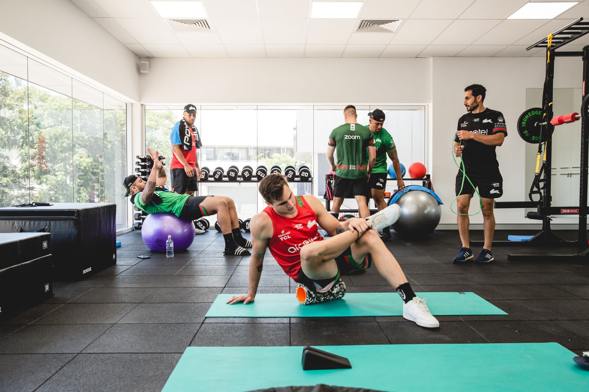 Athletes in a gym doing exercises, with a coach observing. Training area with weights and fitness balls.