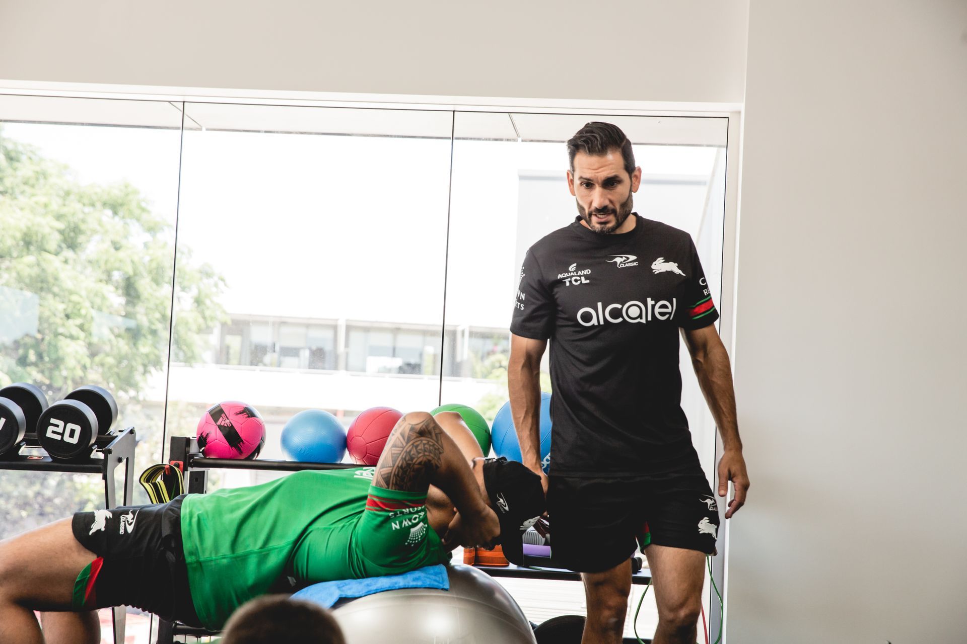 Man supervising another doing crunches on a fitness ball. Indoor gym setting with weights and colorful balls.