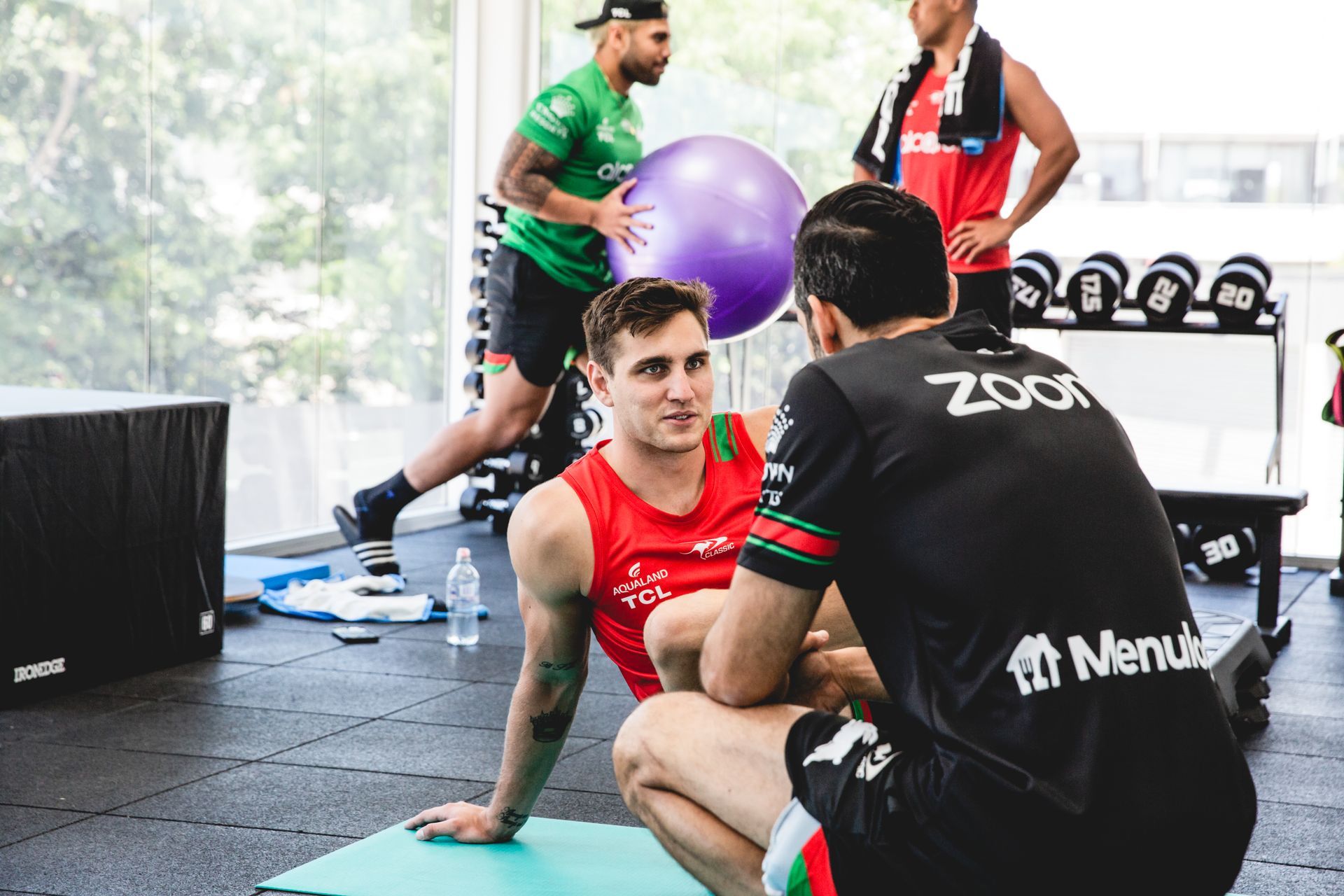 Men in a gym; two conversing, one with ball, others near weights.